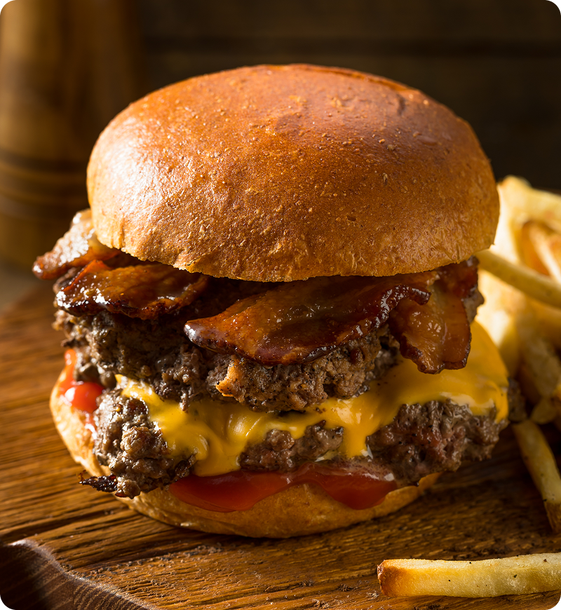 Cheeseburger with bacon, two beef patties, cheddar cheese, and tomato slices on a bun, served with fries on a wooden board.