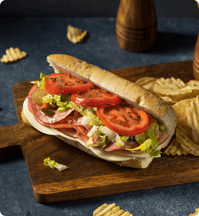 A sub sandwich with layers of meat, cheese, lettuce, and tomato on a wooden board, accompanied by potato chips.