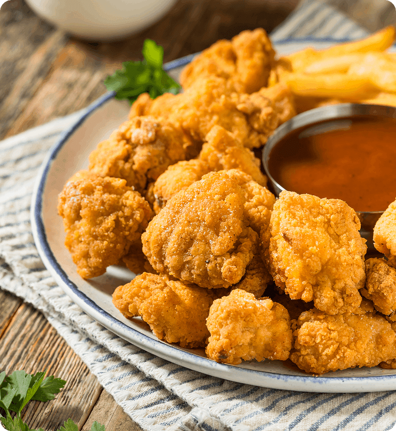 A plate of crispy chicken nuggets served with a small bowl of dipping sauce and a few fries on the side, placed on a wooden table with a striped cloth.
