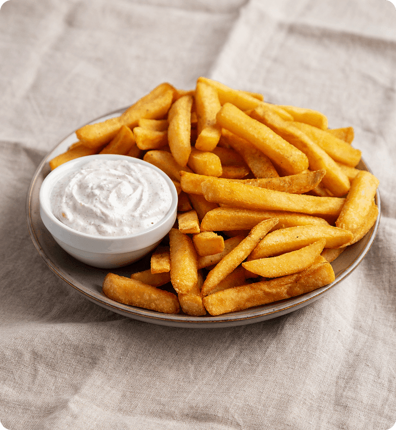 A plate of golden French fries served with a small bowl of creamy white dipping sauce on a light fabric background.