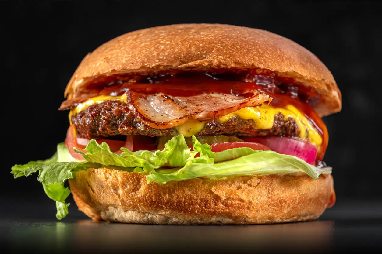 New Project (1) Close-up of a burger with lettuce, tomato, onion, bacon, and melted cheese, topped with sauce and encased in a golden brown bun.